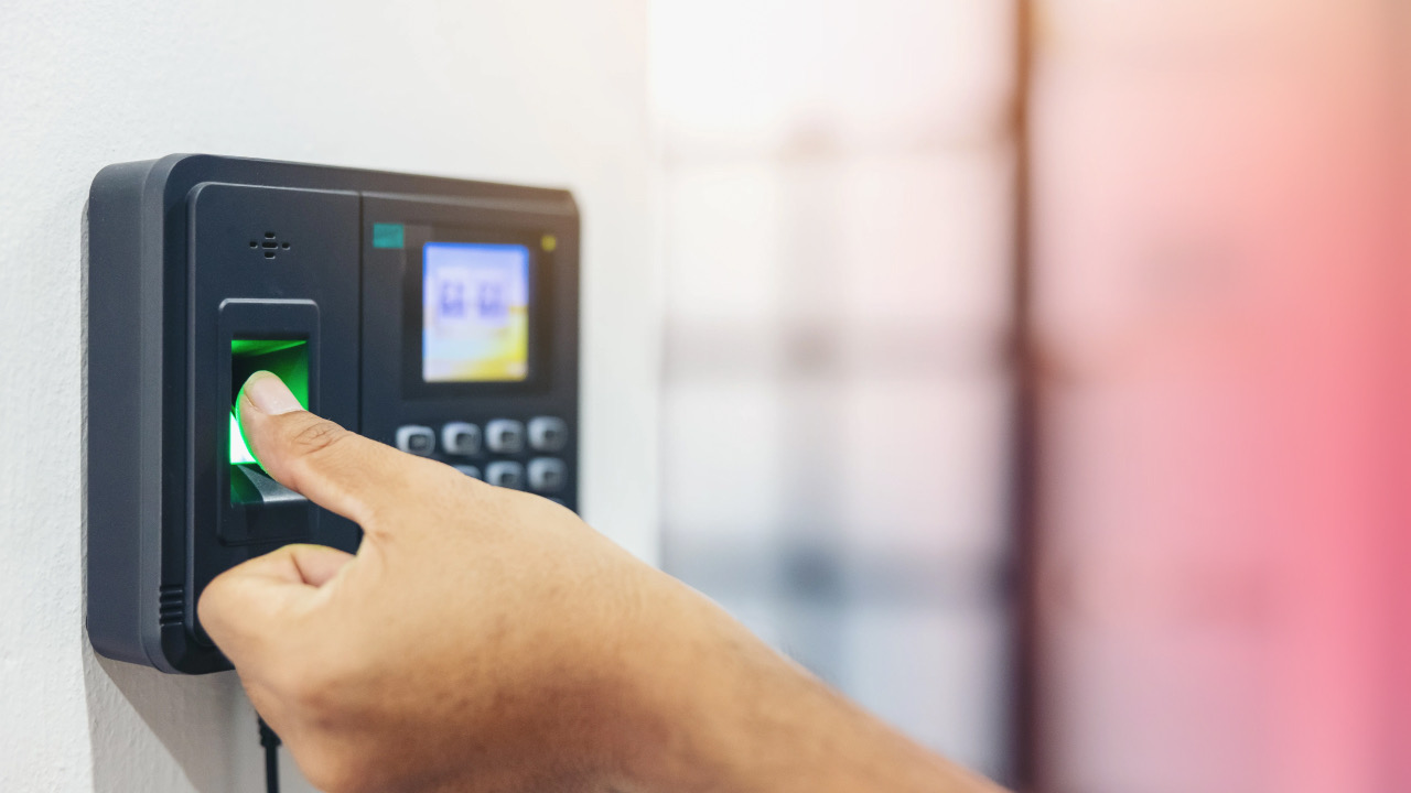 A thumb pressed to the green-lit fingerprint sensor of a wall-mounted time clock with a keypad and small display